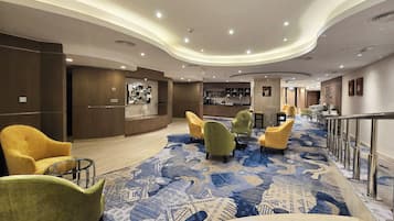 Un hall d’hôtel au design moderne, avec un tapis à motifs, un espace salon avec des fauteuils, un comptoir d’accueil et un bar.