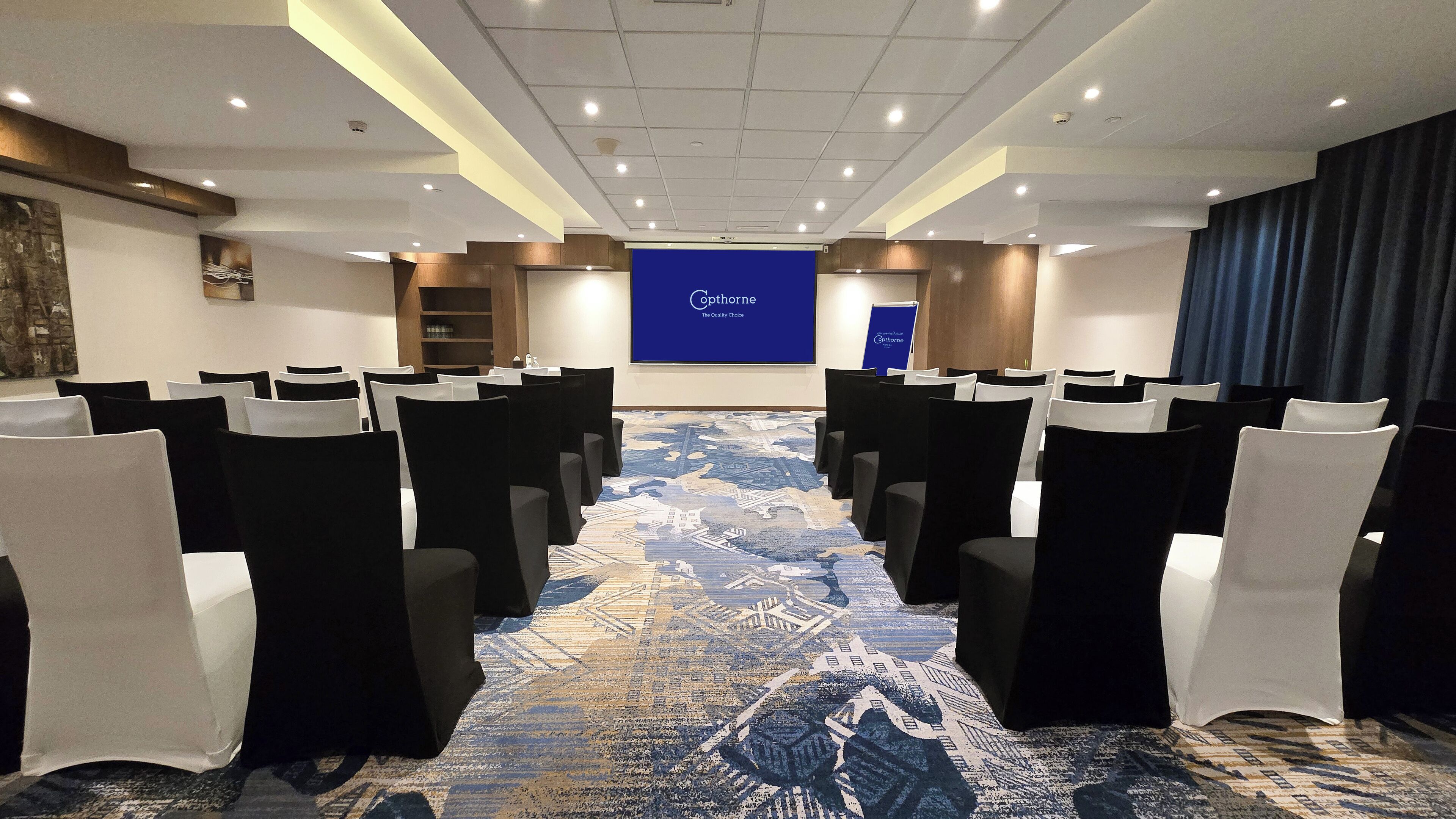 Ein Konferenzraum mit Reihen von Stühlen, einem Teppich mit Stadtblick-Motiv und einer blauen Leinwand mit 'Cophorne'-Branding.
