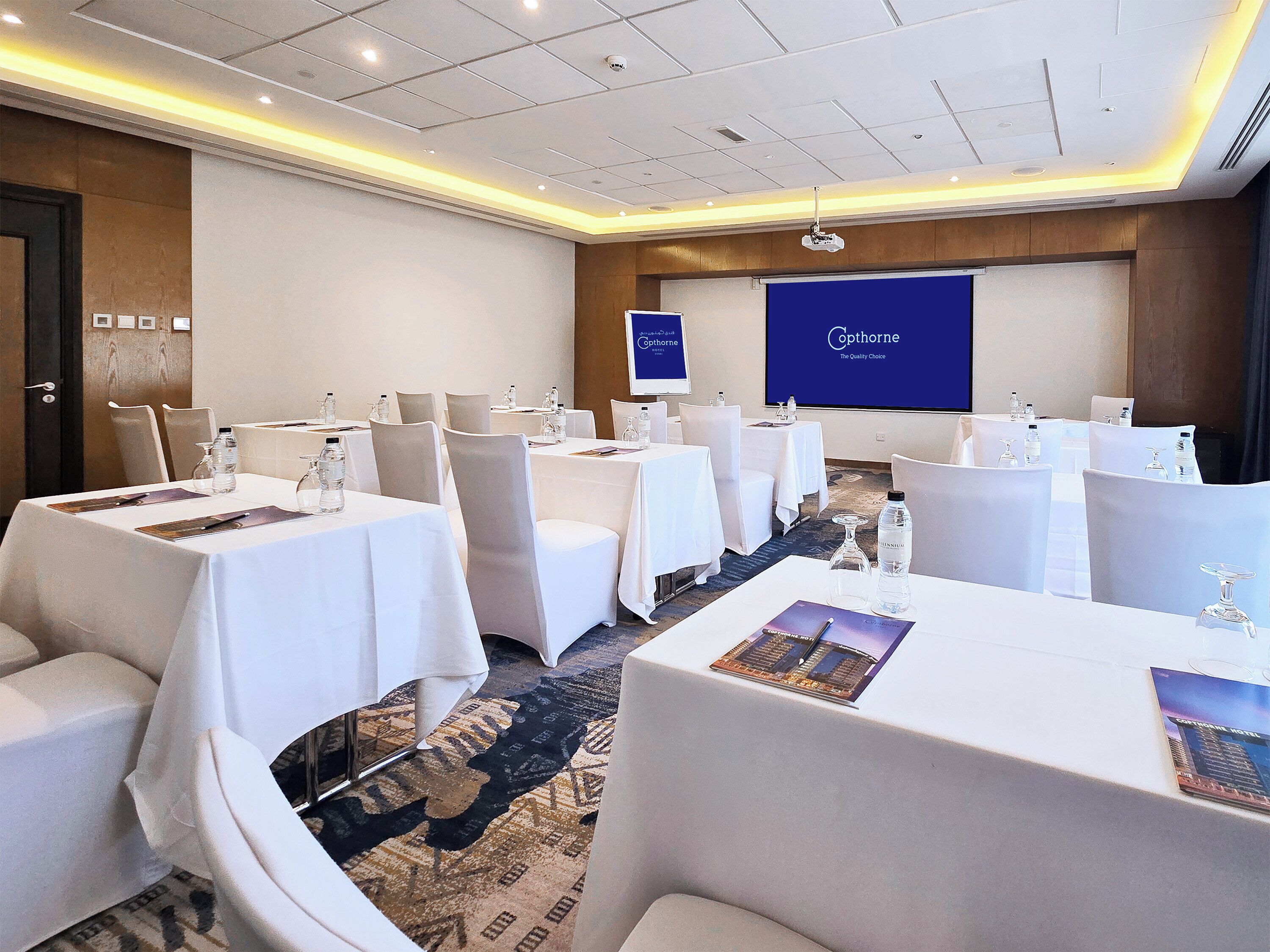 Une salle de conférence équipée de plusieurs tables, de chaises et d’un grand écran affichant le nom de l’hôtel.