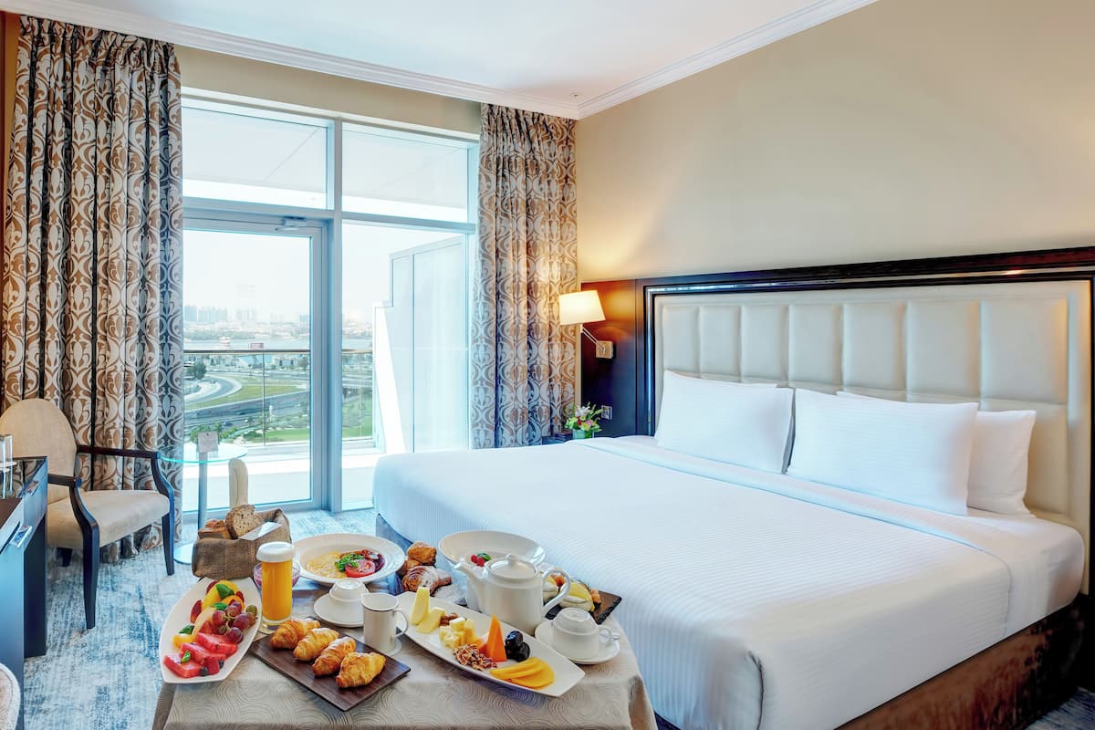 A hotel room with a large bed, a dining table with breakfast items, and a view of a cityscape through the window.