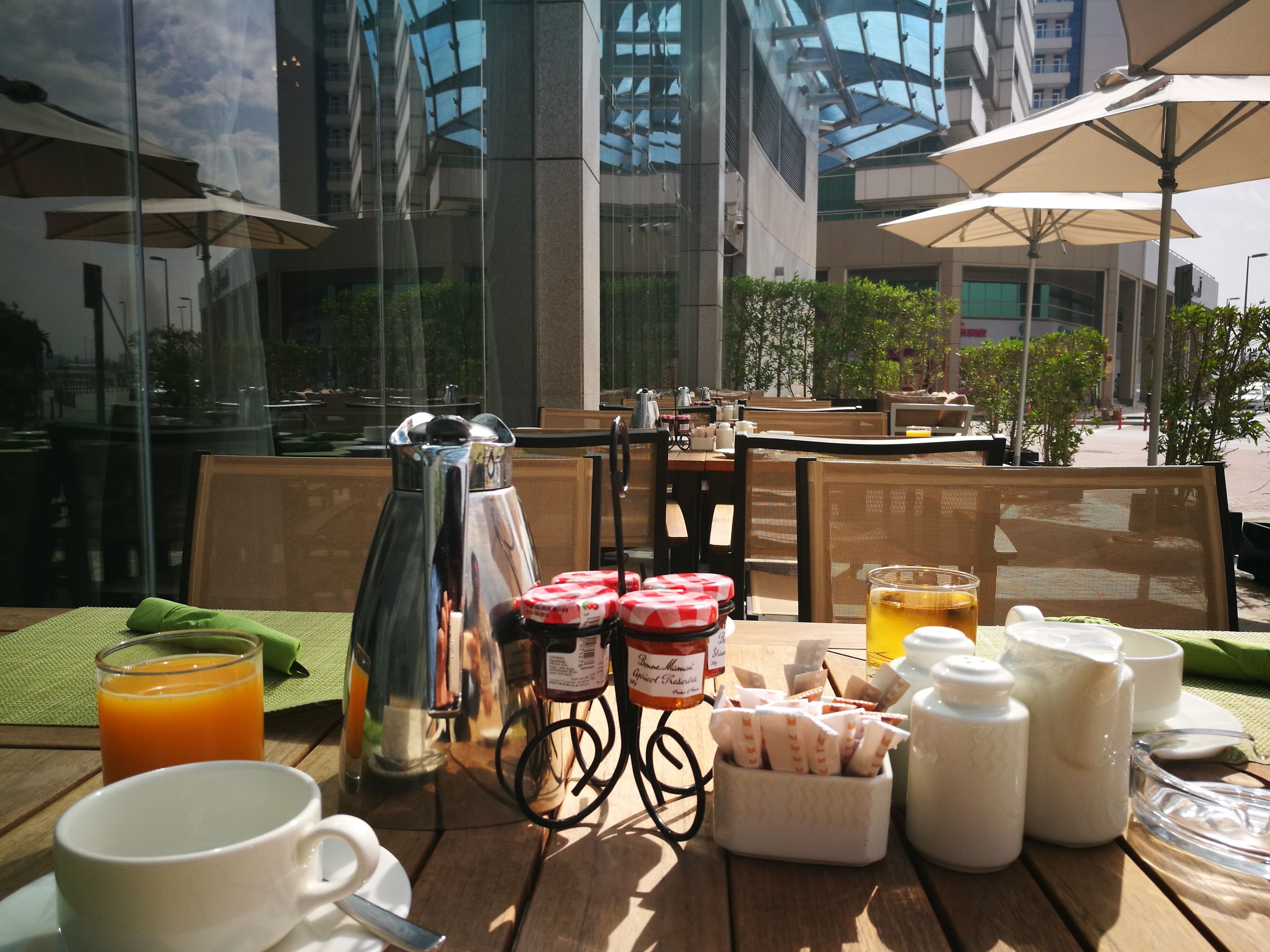 Une table de petit-déjeuner avec une cafetière, du jus, des condiments et une vue sur un bâtiment moderne aux fenêtres en verre.