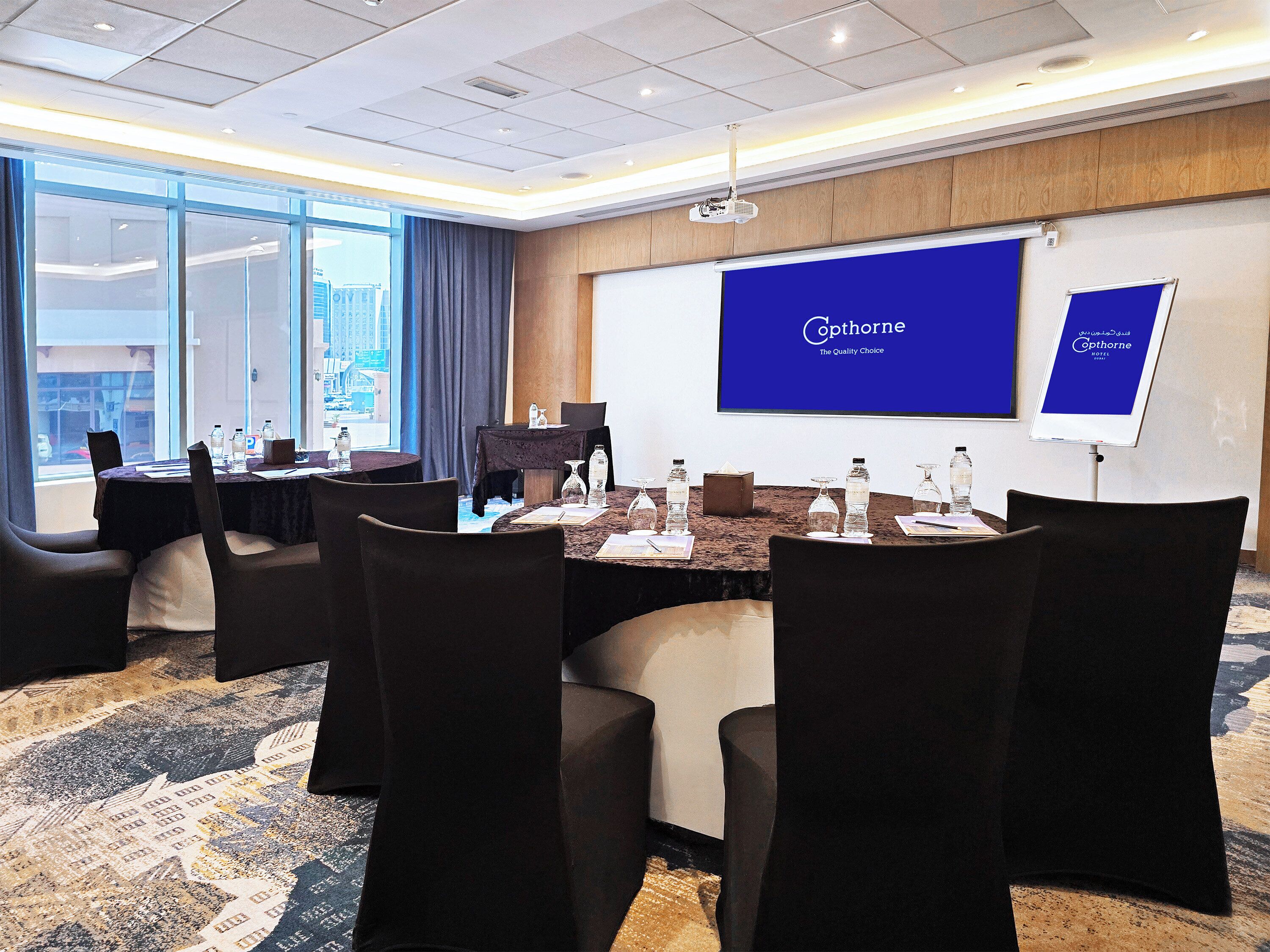 Ein Konferenzraum mit großer Leinwand, die den Hotelnamen zeigt, einem Tisch mit Stühlen und Blick auf die Stadt durch hohe Fenster.