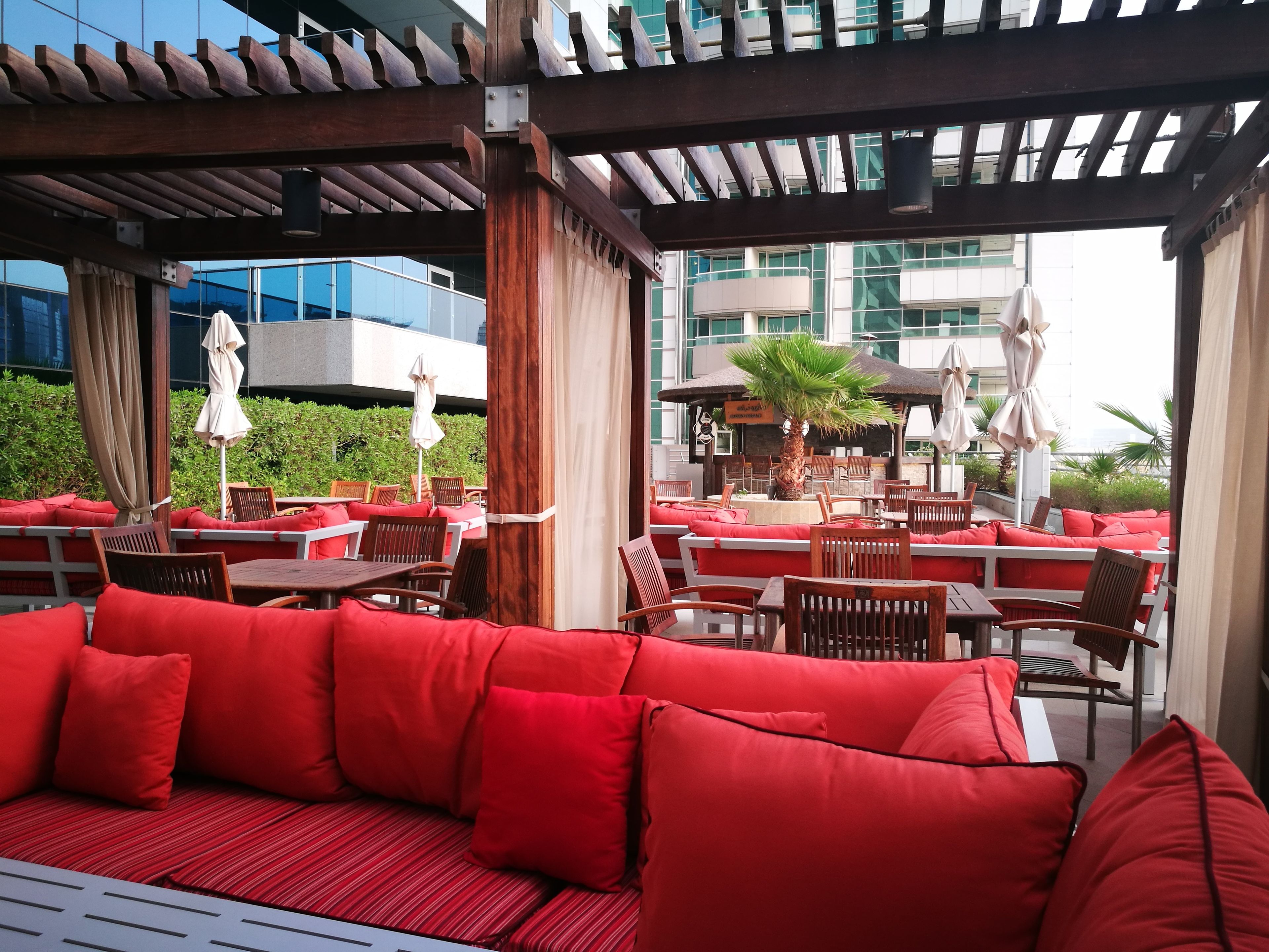 Een terras met rode kussens en houten tafels, met uitzicht op een modern gebouw met grote ramen.