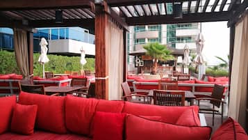An outdoor seating area with red cushions and wooden tables, overlooking a modern building with large windows.