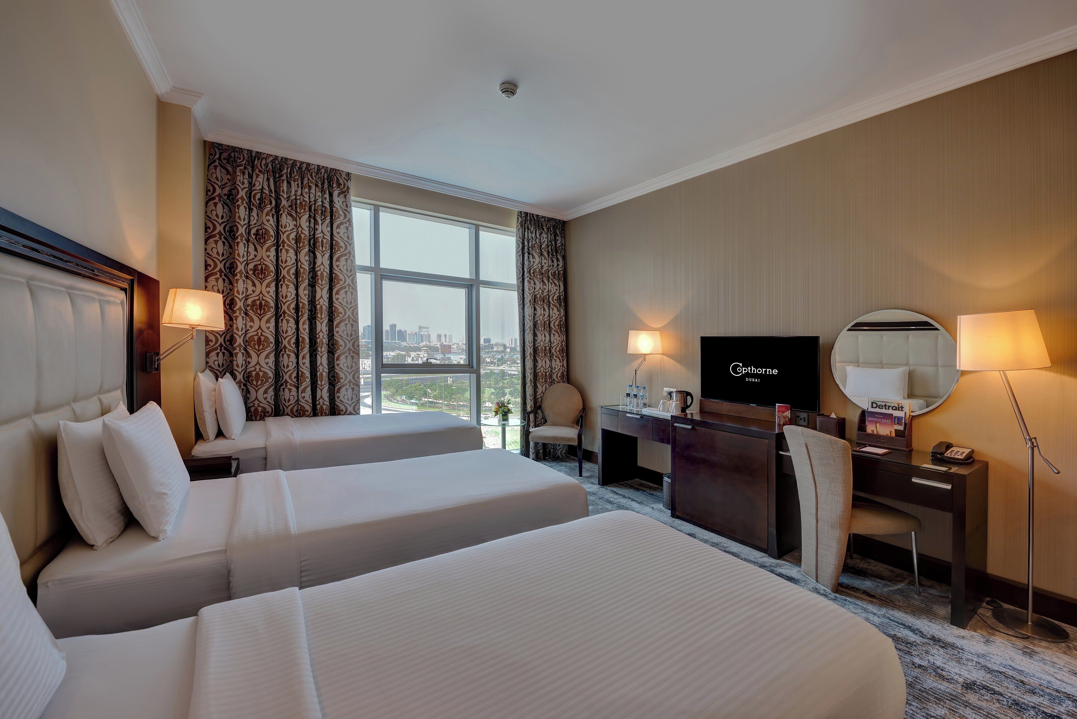 A hotel room with two beds, a desk, a chair, a TV, and a view of the city.