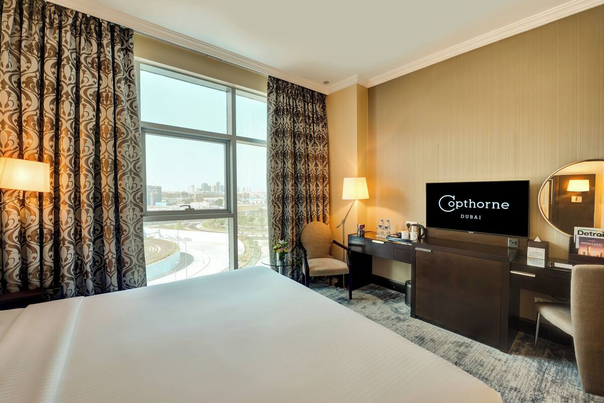A hotel room with a large bed, a desk with a flat-screen TV, a chair, a desk lamp, and a view of the city.