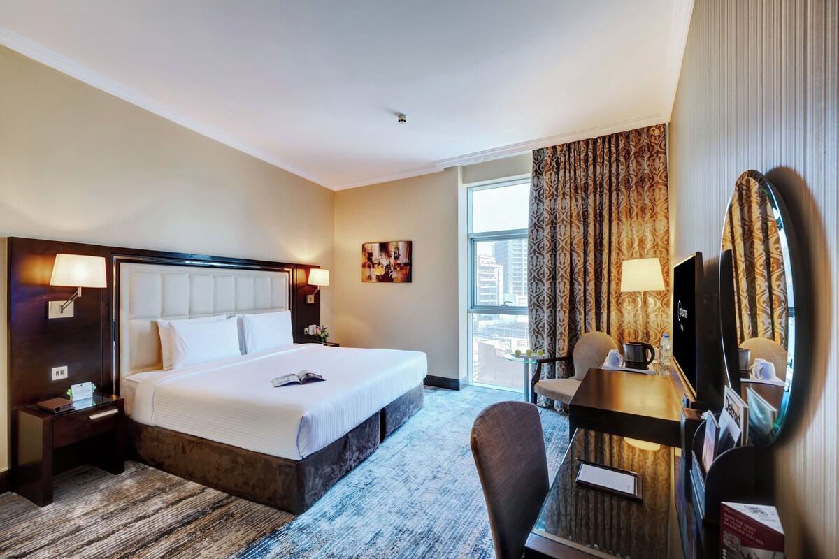 A hotel room with a large bed, a desk with a chair, a television, and a window with curtains.