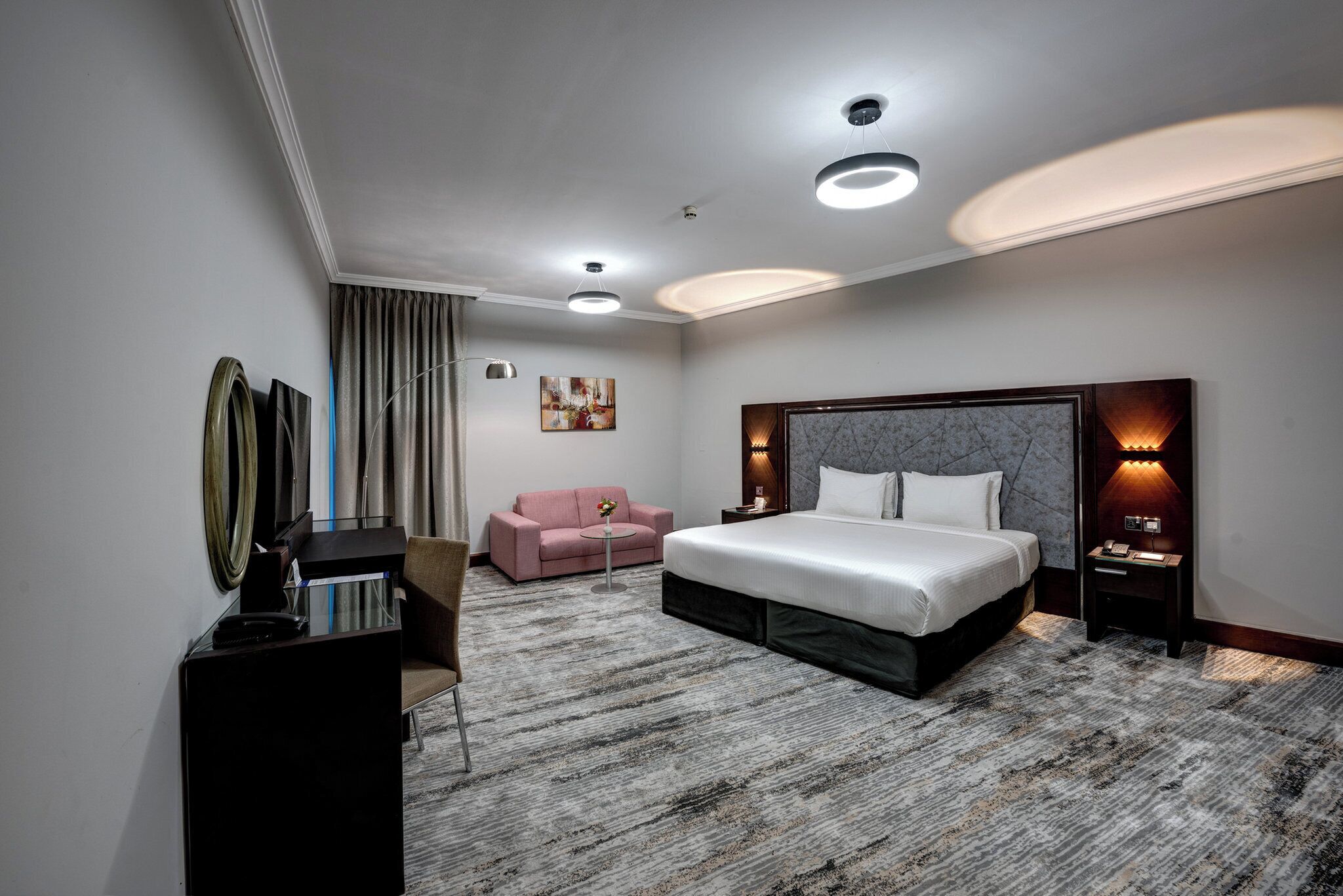 A modern hotel room with a large bed, a desk, a pink sofa, and a small table.