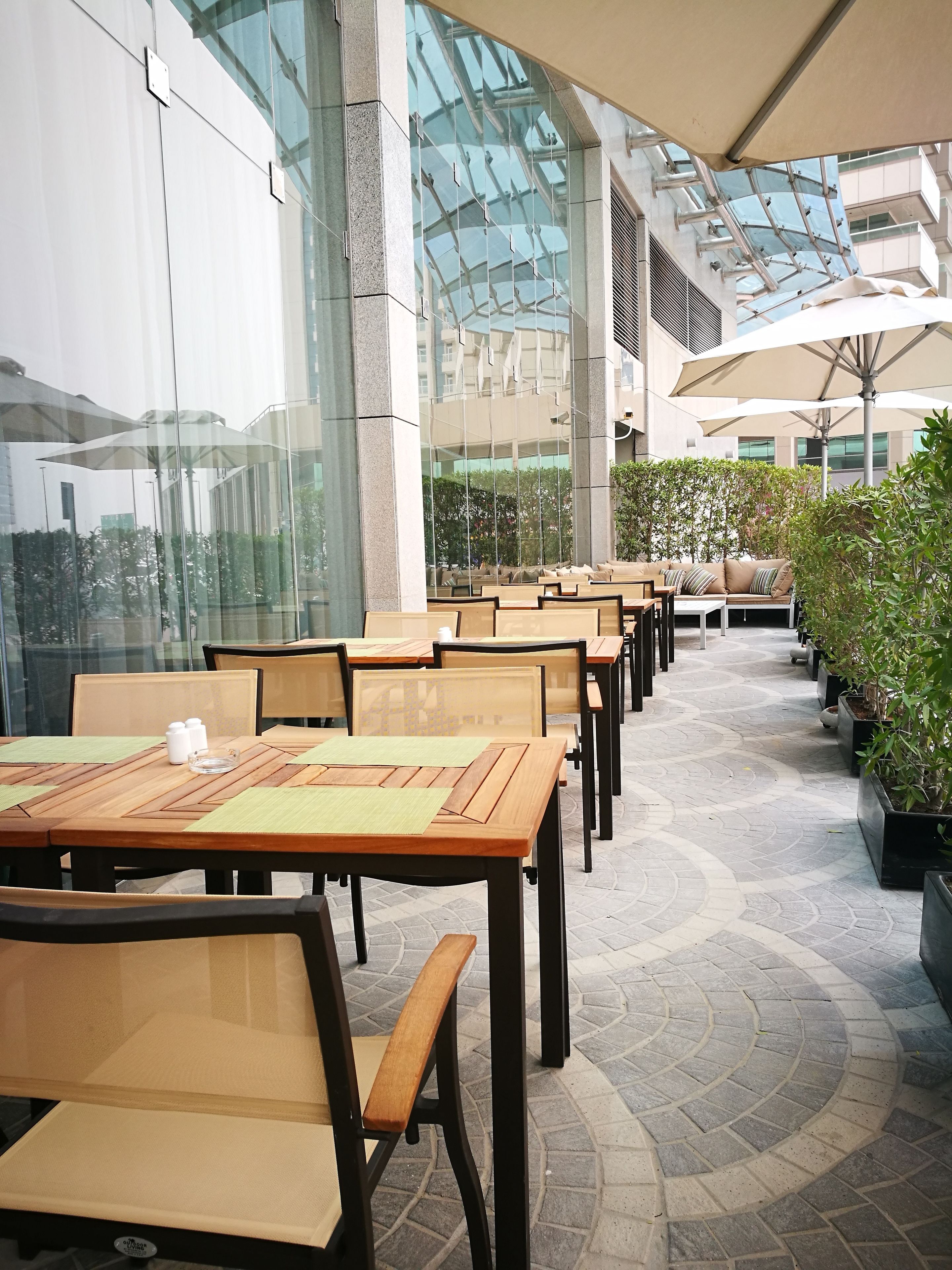An outdoor cafe area with wooden tables and chairs, large umbrellas, and potted plants.