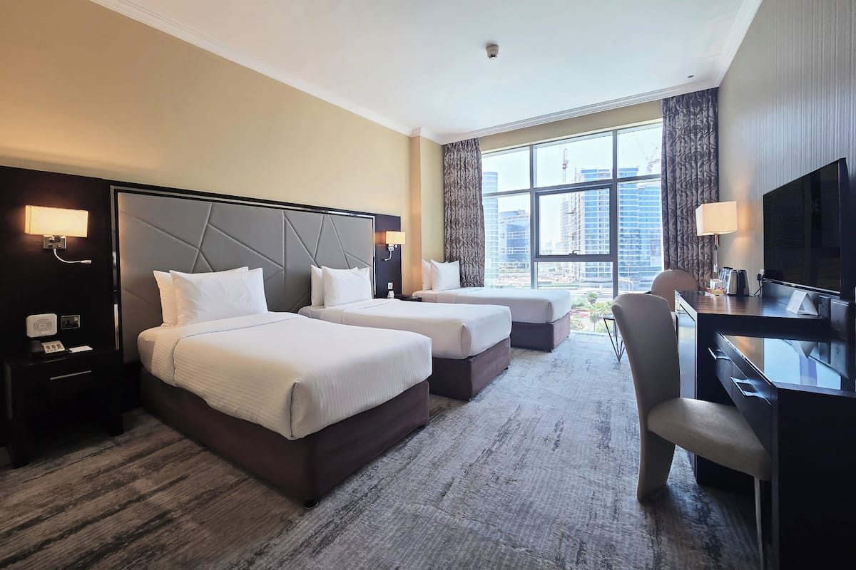 A hotel room with two beds, a desk, a chair, a TV, and a large window with a city view.