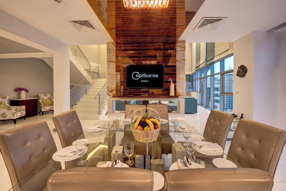 A dining area with a glass table, leather chairs, and a fruit basket. A television screen displays "Coptorne Dubai".