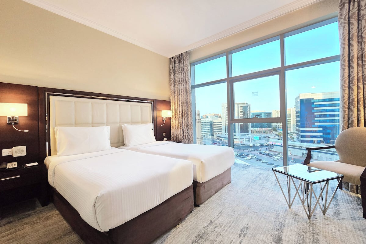 A hotel room with two beds, a large window with a city view, a small table, and a chair.