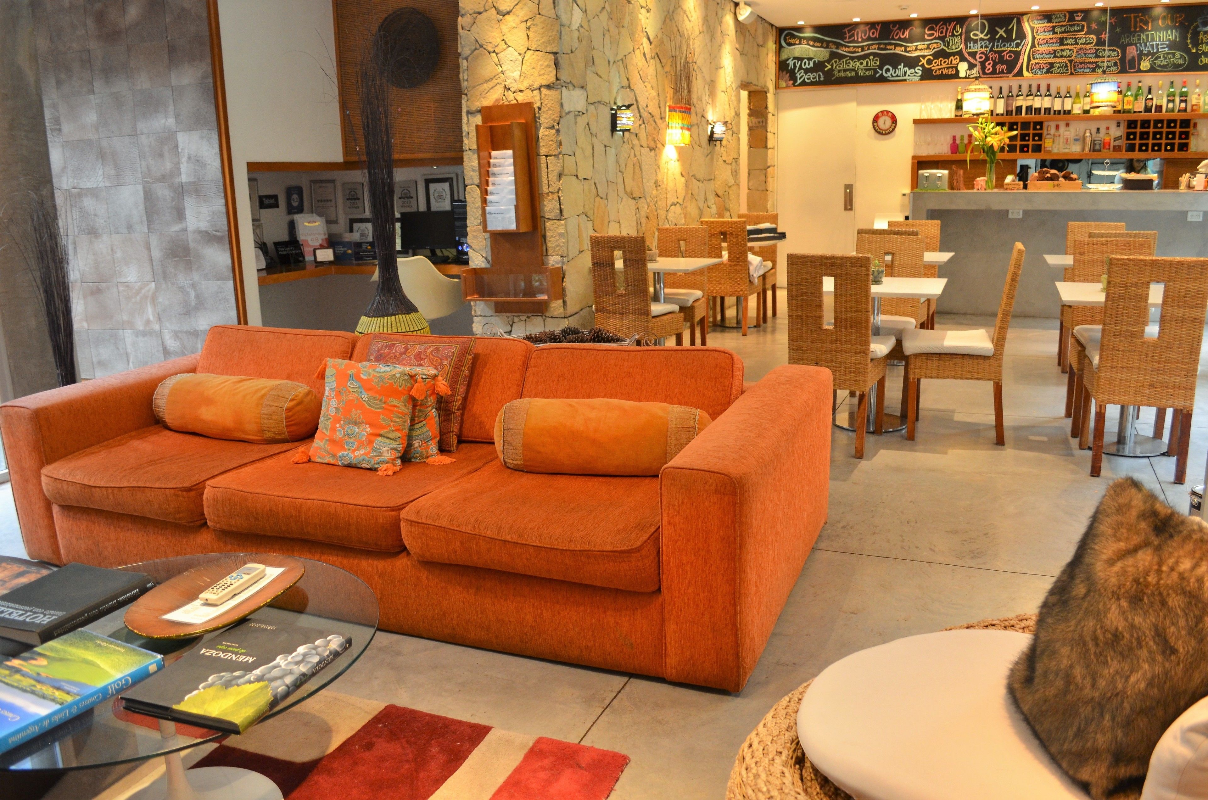 A hotel lobby with a large orange sofa, wicker chairs, a bar area with a wine selection, and a stone accent wall.