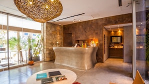Un área de recepción moderna con pared de piedra, un mostrador de recepción, una mesa redonda de café con libros, un tapete rojo y una silla de mimbre con cojín amarillo.