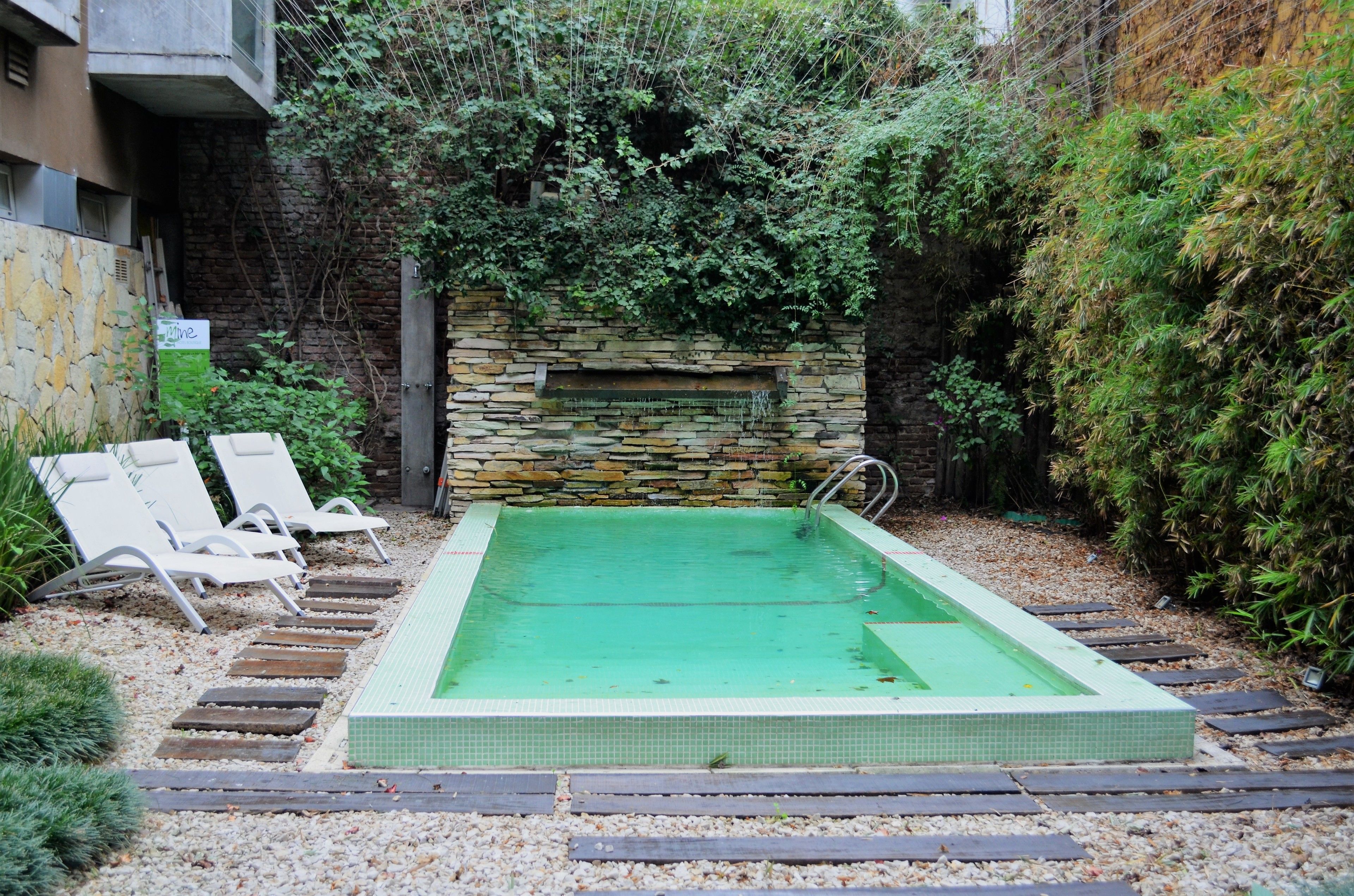 A rectangular pool with a stone wall and a small waterfall feature, surrounded by a pebble path and two white lounge chairs.