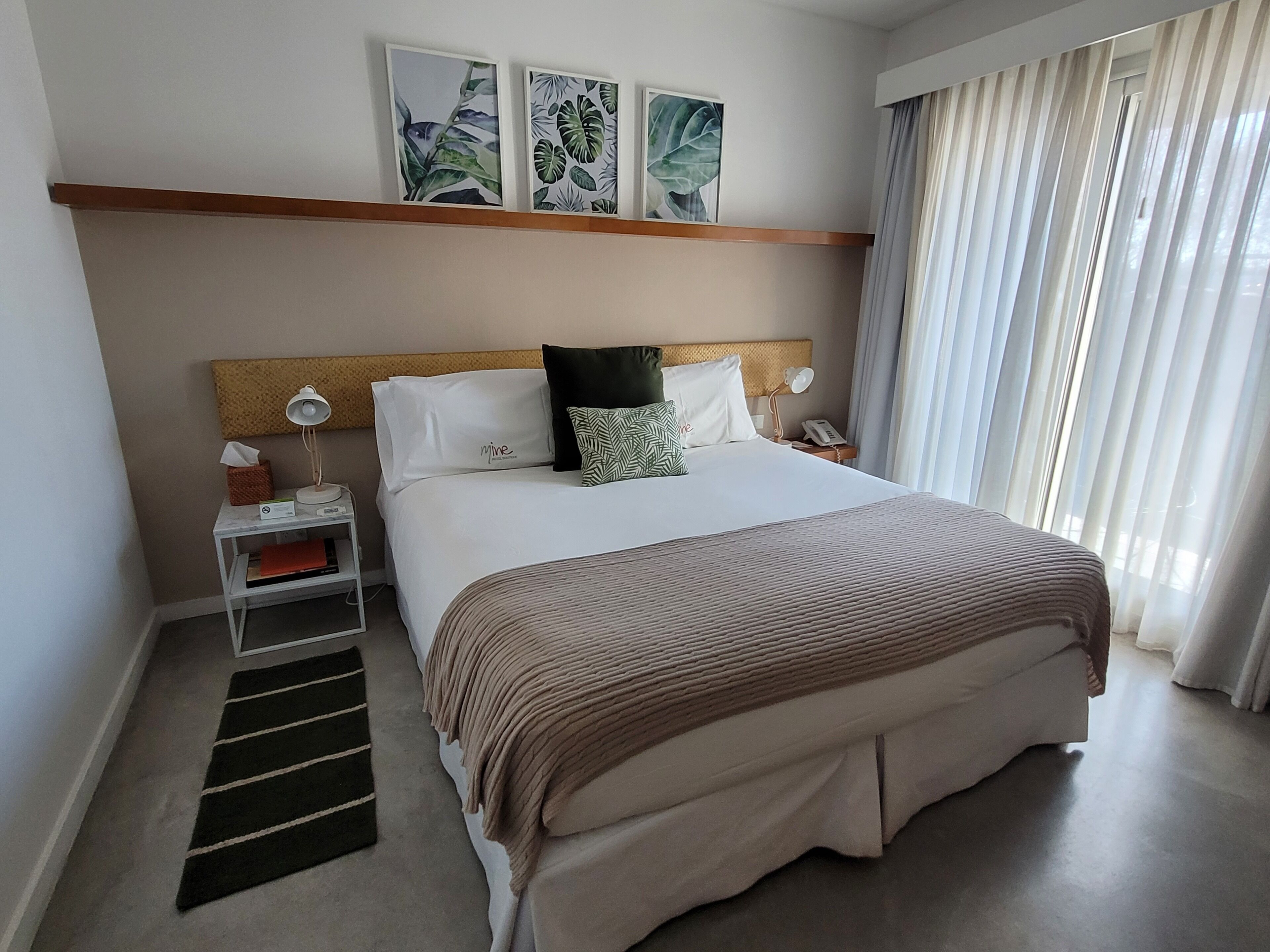 A hotel room with a large bed, a bedside table with a lamp, a small shelf with a tissue box, and a window with curtains.
