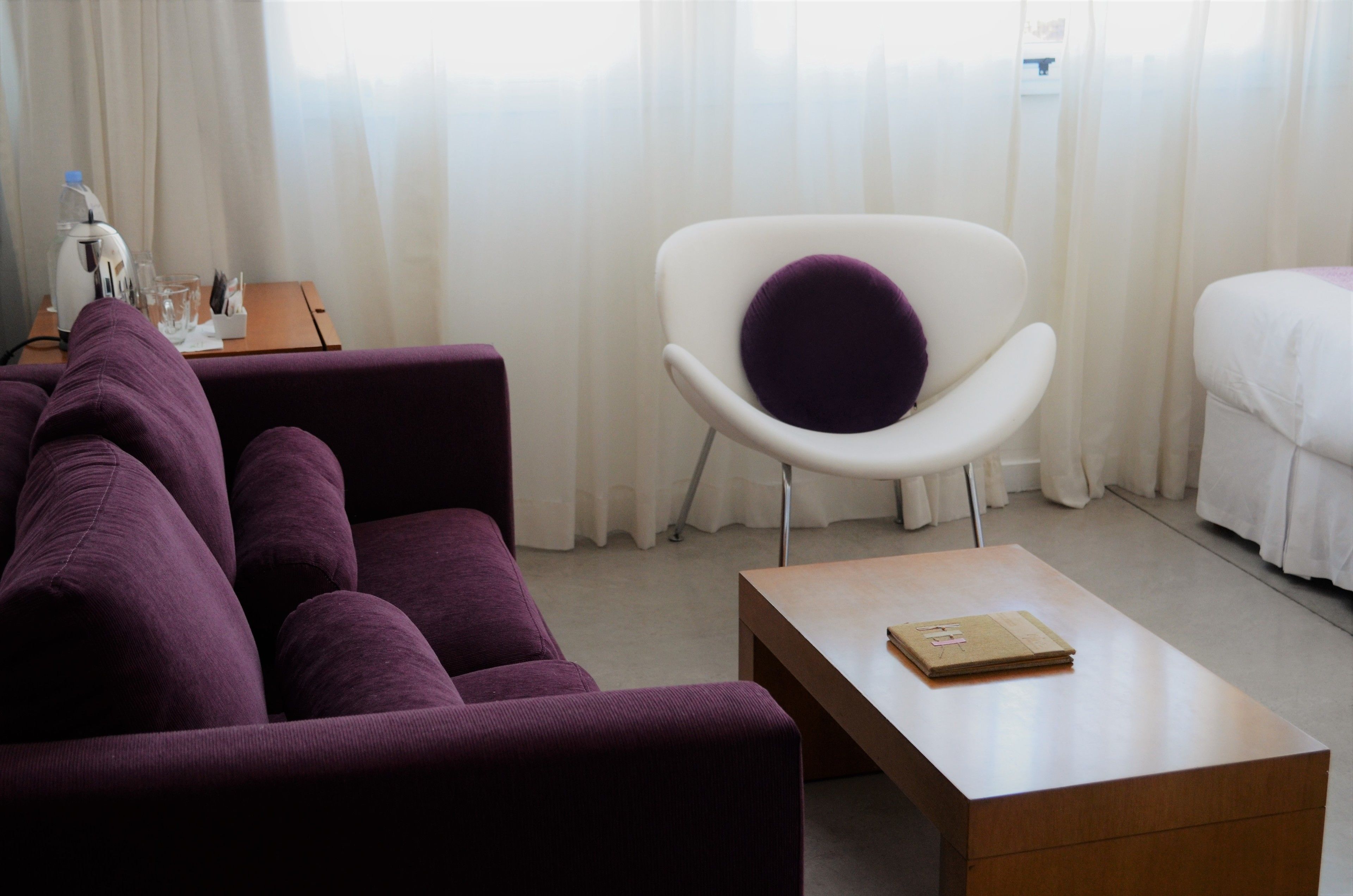 A modern hotel room with a purple sofa, a white armchair with a purple cushion, a wooden coffee table, and a bed with white bedding.