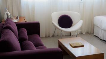 A modern hotel room with a purple sofa, a white armchair with a purple cushion, a wooden coffee table, and a bed with white bedding.