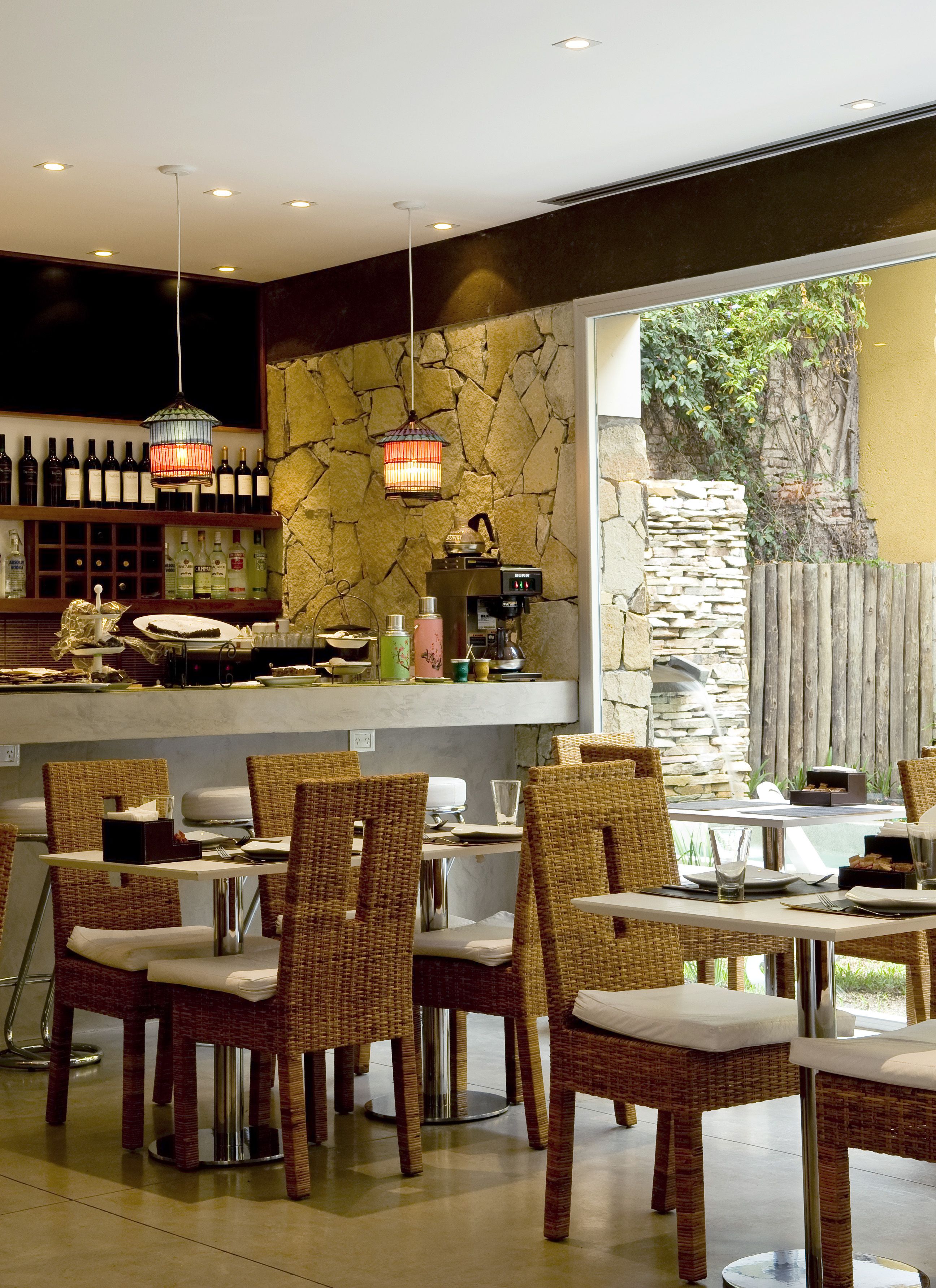 A restaurant with wicker chairs, a bar area with wine bottles, and a stone wall.