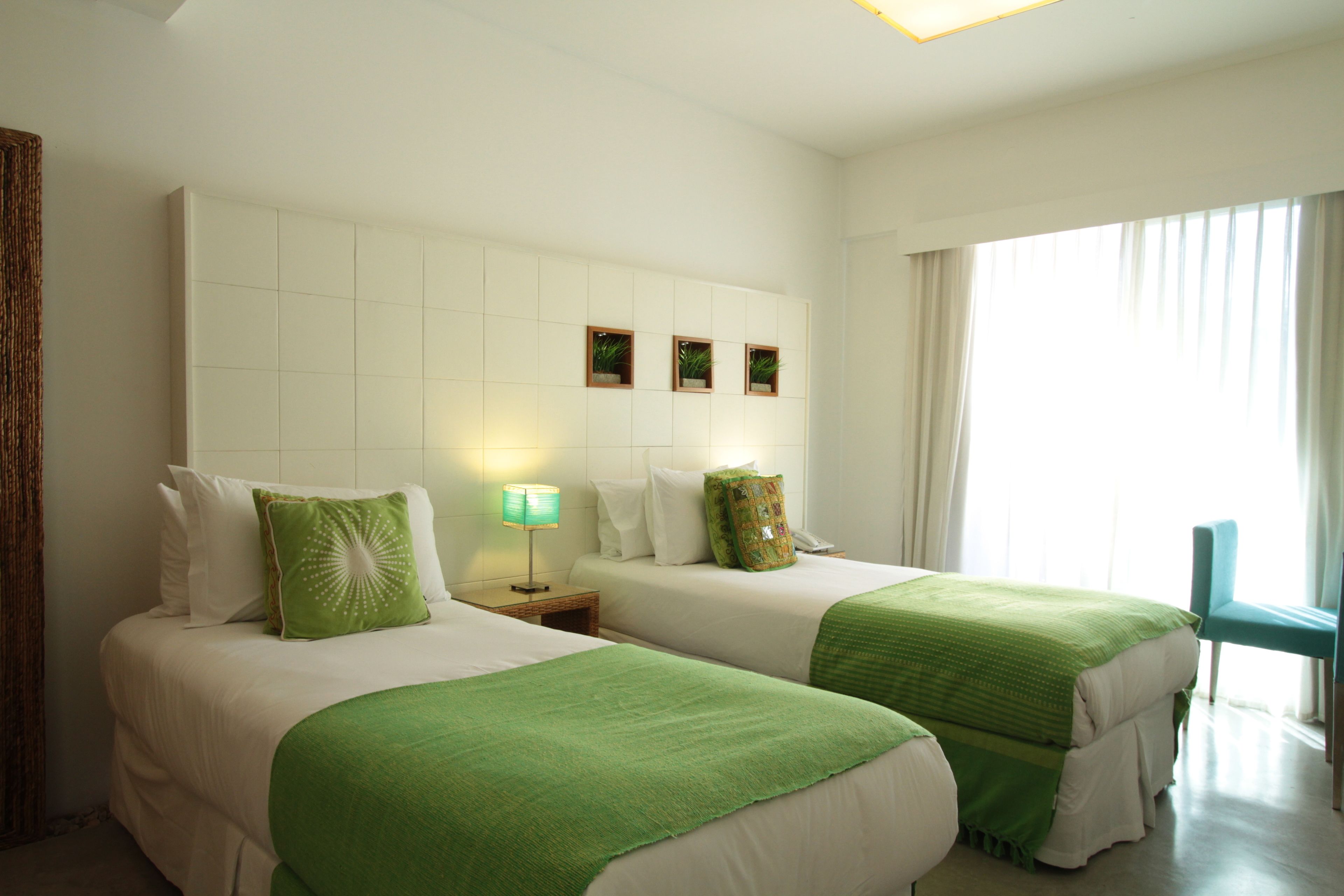 A hotel room with two beds, a nightstand with a lamp, a window with curtains, and a wall with three small framed pictures.