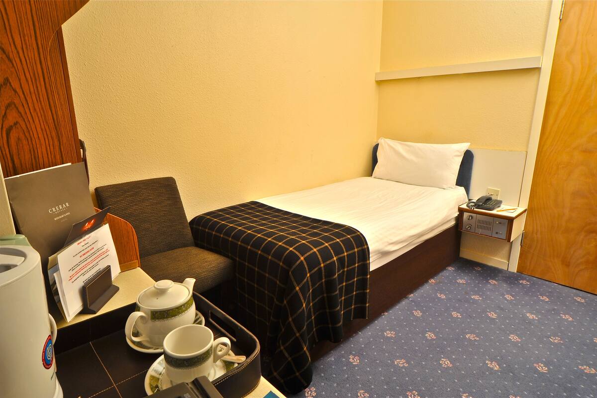Ein Hotelzimmer mit Bett, Nachttisch mit Lampe, Sessel, kleiner Tisch mit Teekanne und Tassen sowie einem Schild mit Zimmerinformationen.