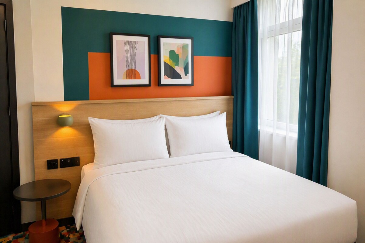 Ein Hotelzimmer mit einem Bett, einem Kopfteil, zwei gerahmten Kunstwerken, einem Nachttisch und einem Fenster mit Vorhängen.