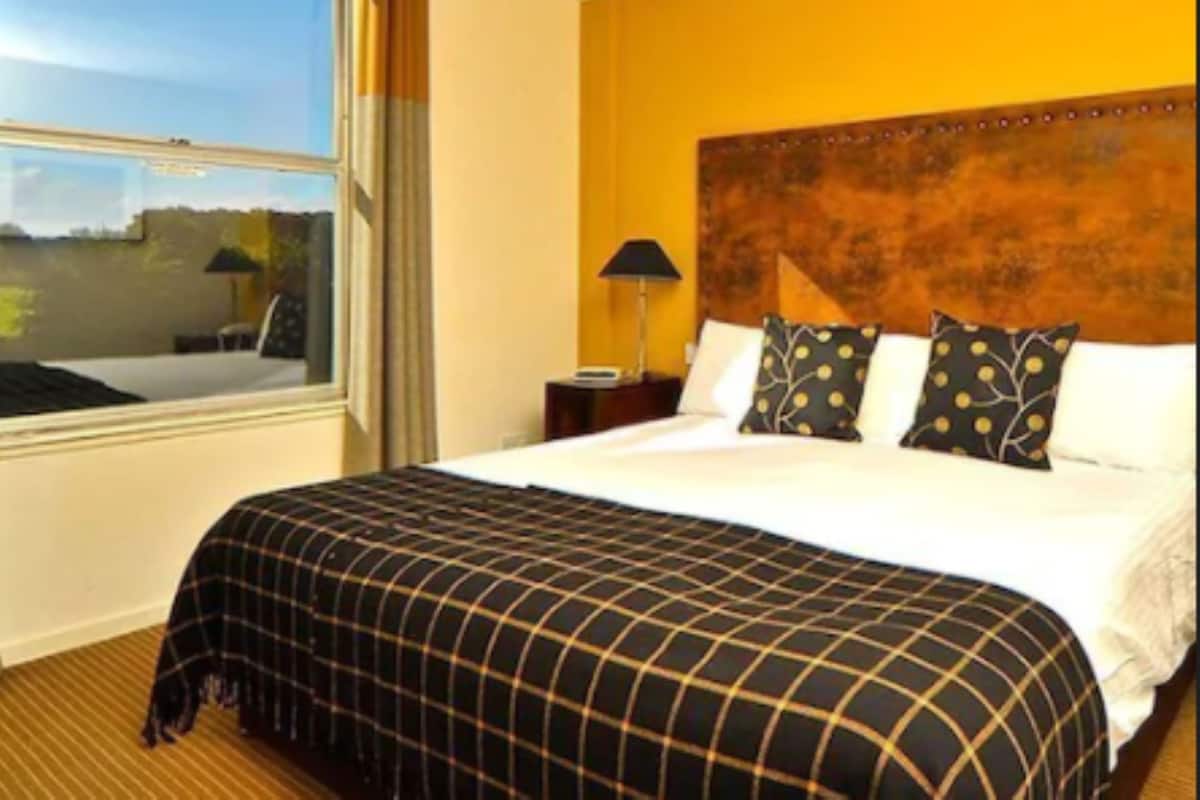 Ein Hotelzimmer mit großem Fenster, einem Bett mit schwarz-weiß kariertem Überwurf und einem hölzernen Kopfteil.