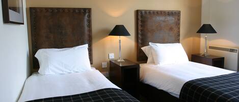 Habitación de hotel con dos camas, cada una con cabecero, mesitas de noche, lámparas y una colcha blanca.