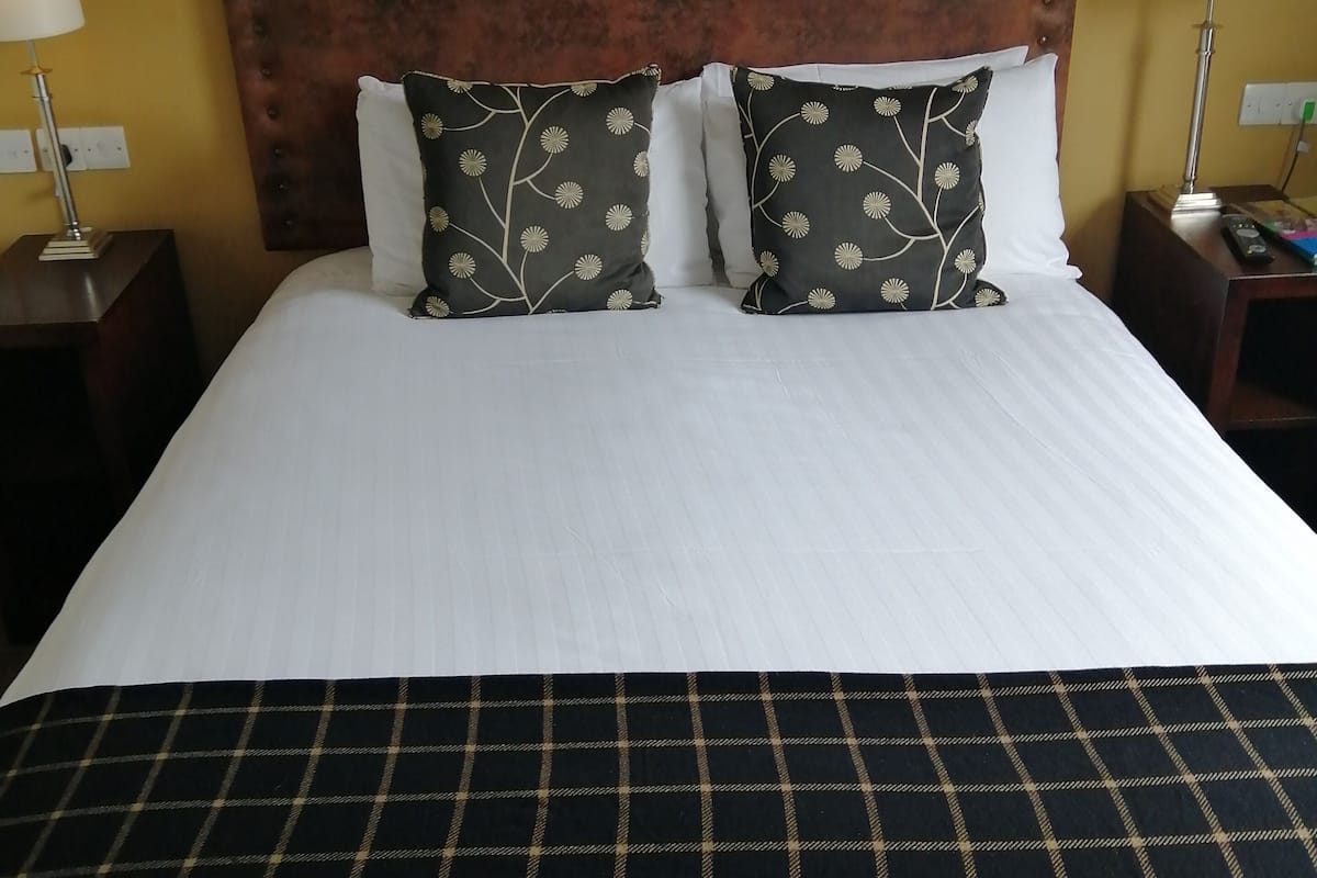Ein ordentlich bezogenes Bett mit einem dunkel karierten Überwurf, zwei Kissen mit Blumenmuster und ein Holzkopfschild.