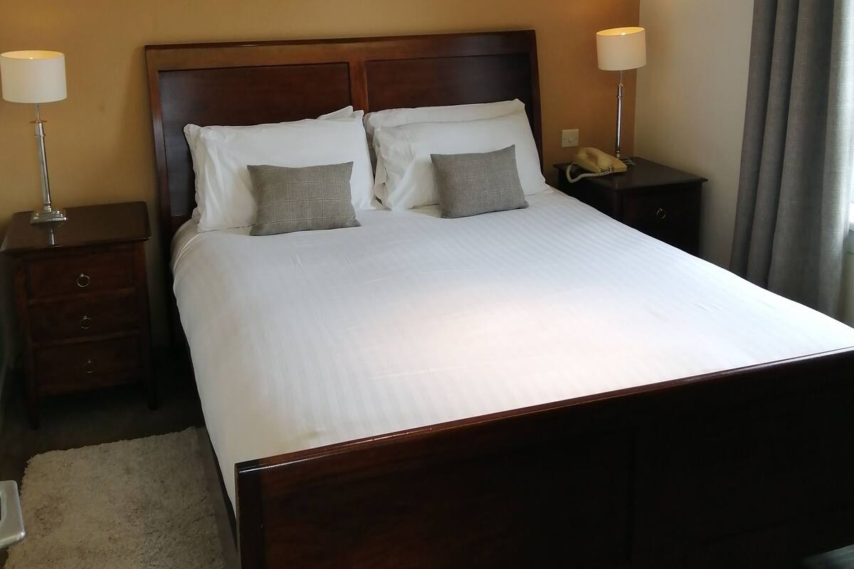Ein Hotelzimmer mit einem großen Bett, zwei Nachttischen mit Lampen, einem Telefon und einem Fenster mit Vorhängen.