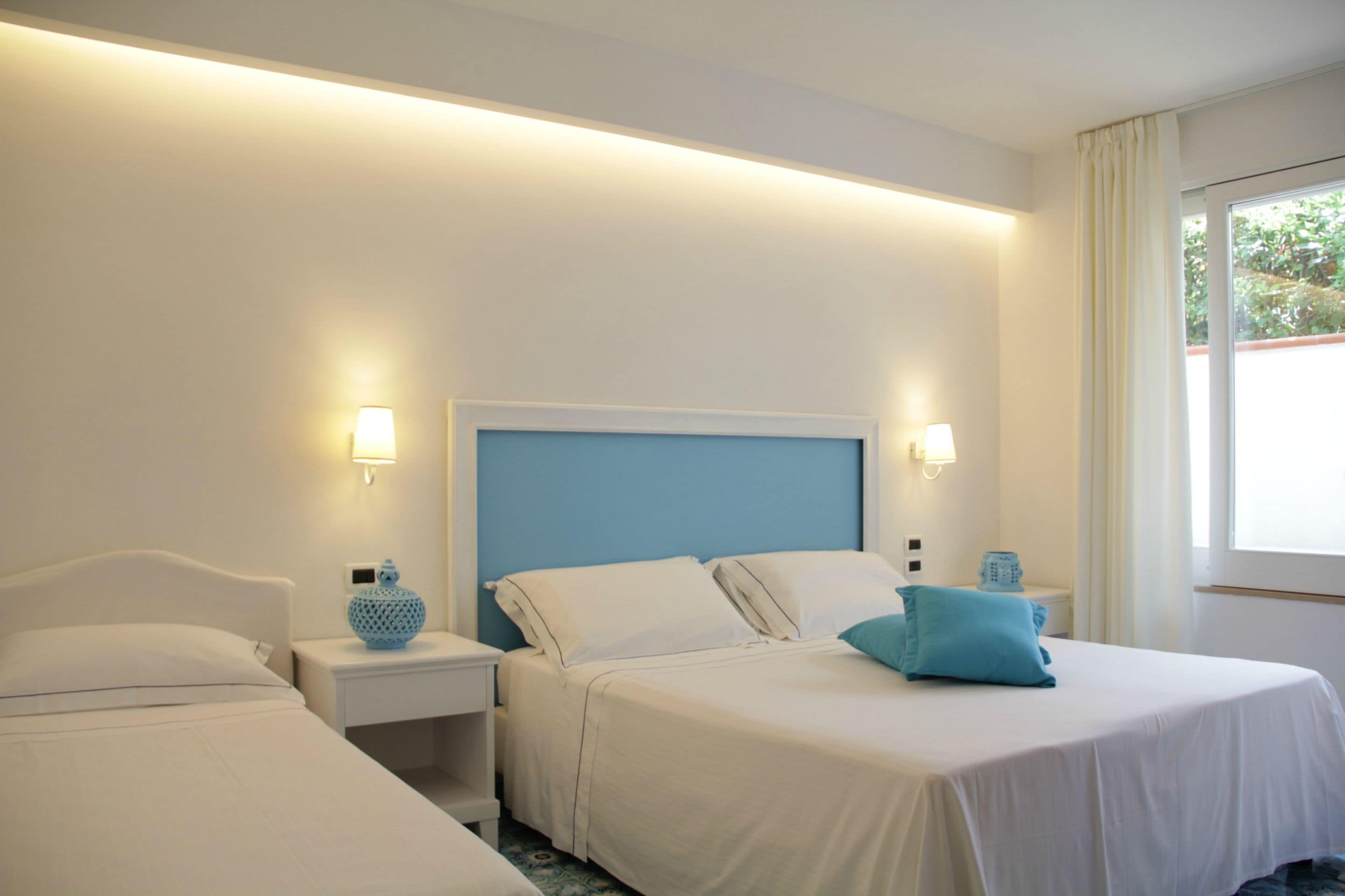 A hotel room with two beds, a blue headboard, bedside lamps, and a window with curtains.