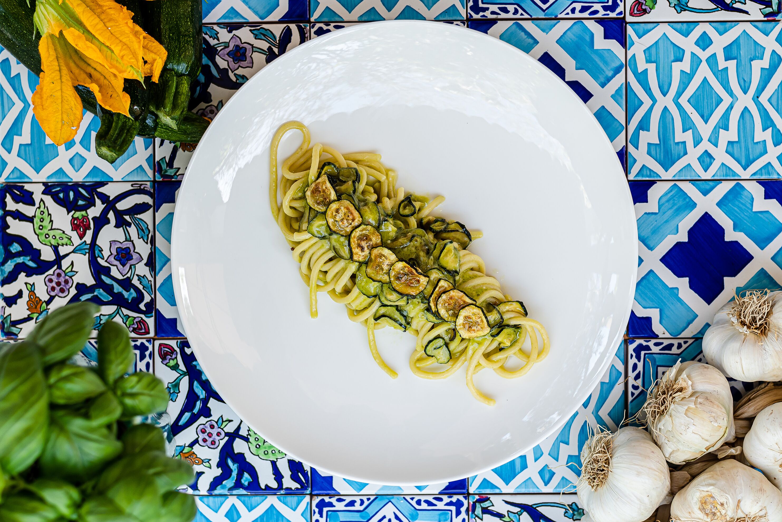 Een bord pasta met groenten, omgeven door knoflookbollen en een basilicplant.