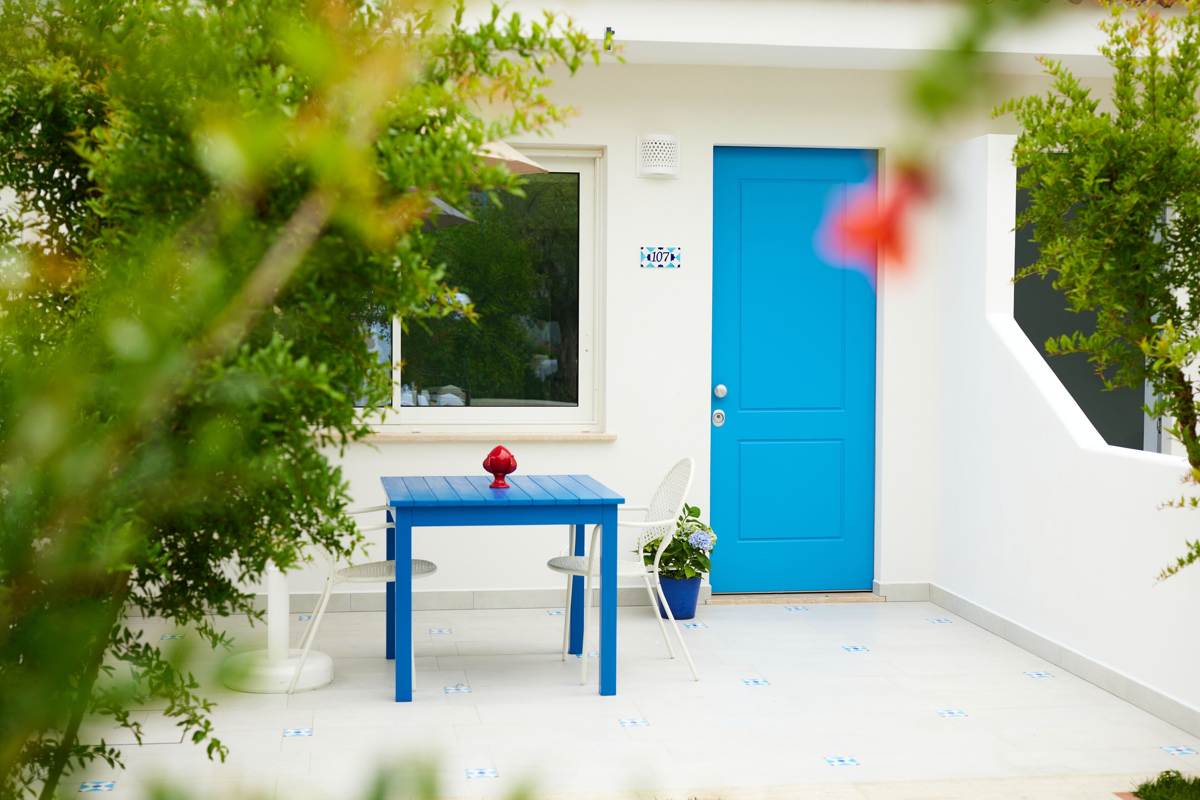 Fachada branca com uma porta azul, uma pequena mesa e um objeto vermelho, além de um vaso com planta.