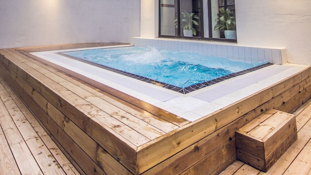 A modern outdoor hot tub area with wooden decking and a pool, surrounded by a building with large windows and wall-mounted lights. - Braavo Spa Hotel (Tallinn)