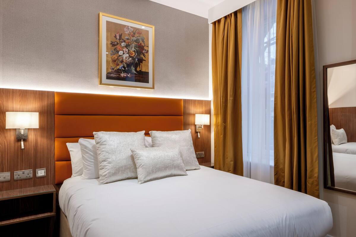 A hotel room with a large bed, two bedside lamps, a framed floral artwork, and a window with curtains.