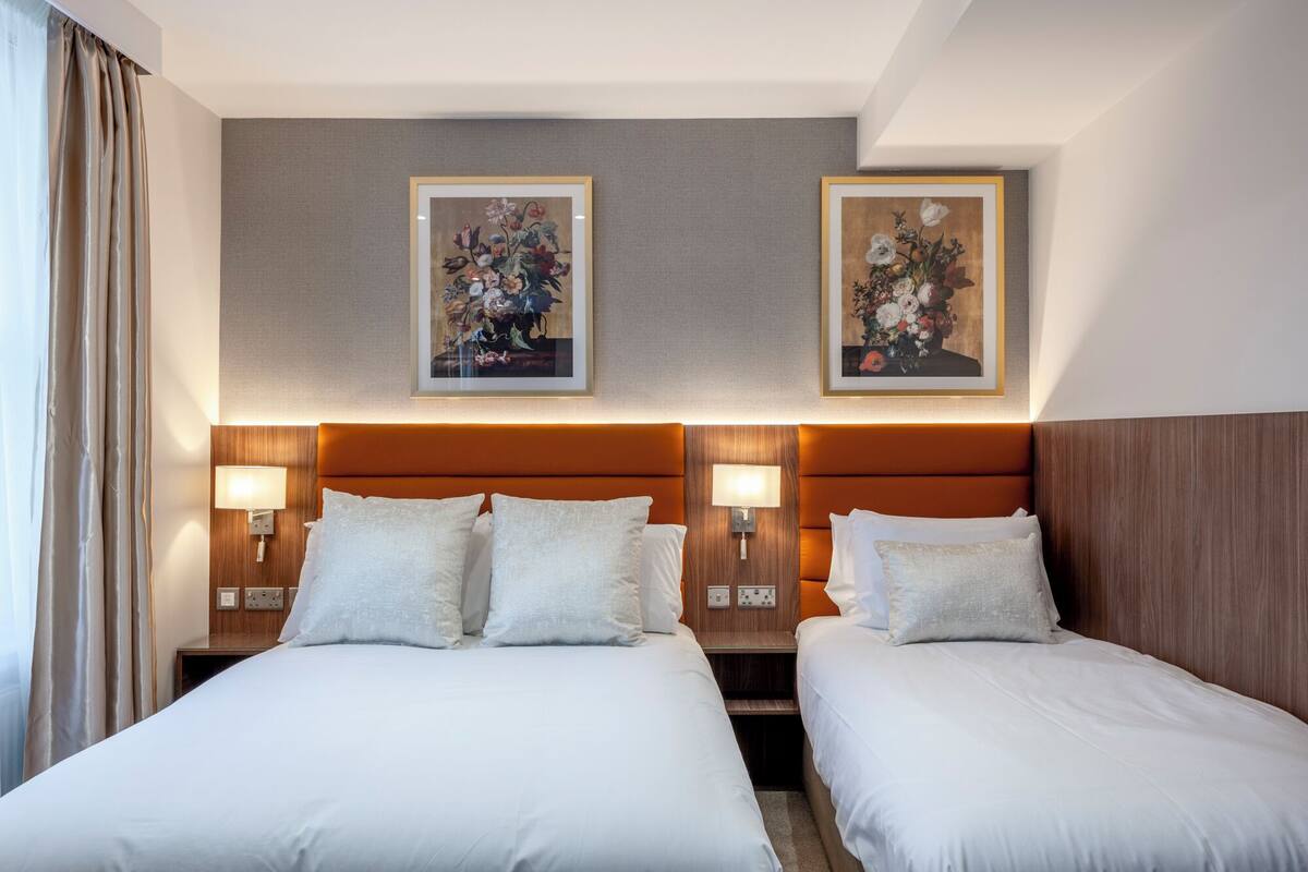 A hotel room with two beds, each with a wooden headboard and bedside lamps. There are two framed floral paintings on the wall above the beds.