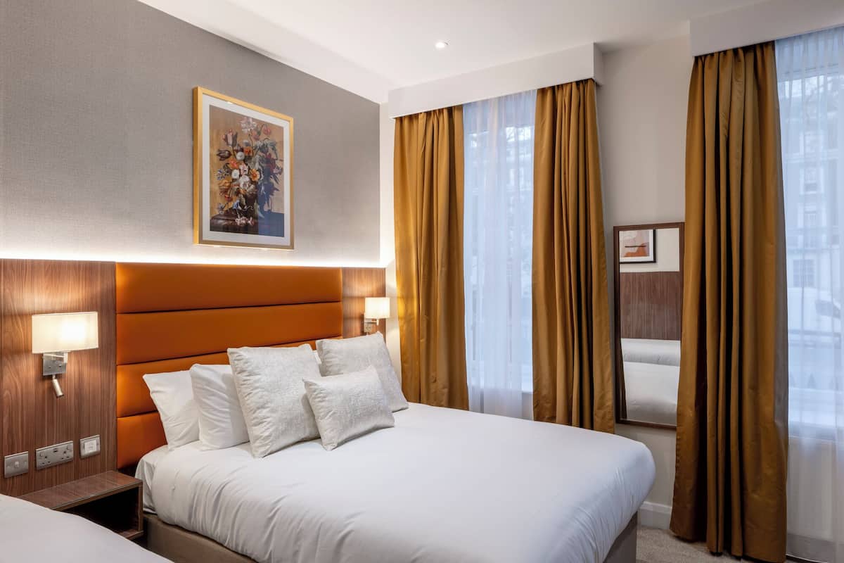 A hotel room with a large bed, a headboard, a bedside table with a lamp, a wall-mounted painting, and a window with curtains.