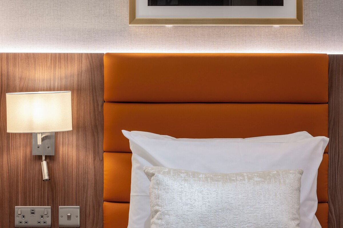 Una habitació d'hotel amb un capçal de llit de fusta, una làmpada de nit, una obra d'art floral emmarcada i un almofí blanc.