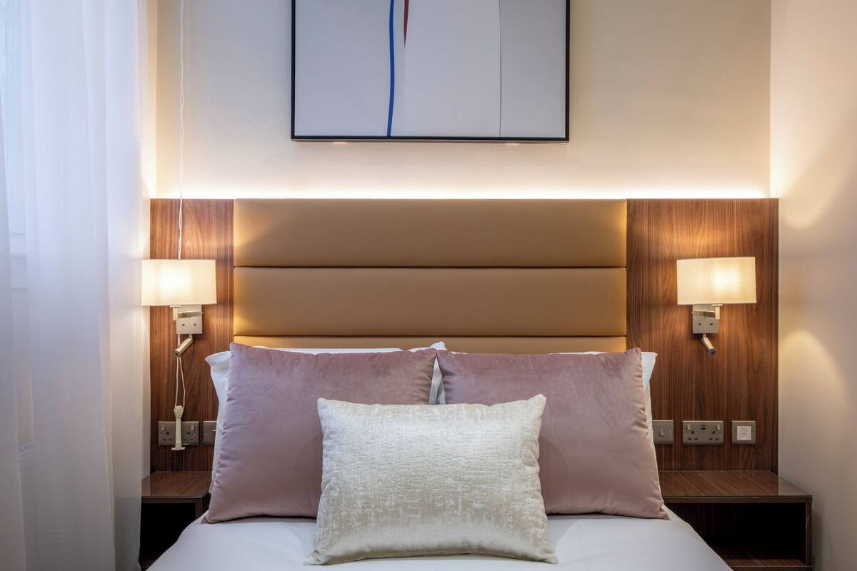 A hotel room with a bed, two bedside lamps, a wall-mounted light, and a framed picture above the bed.