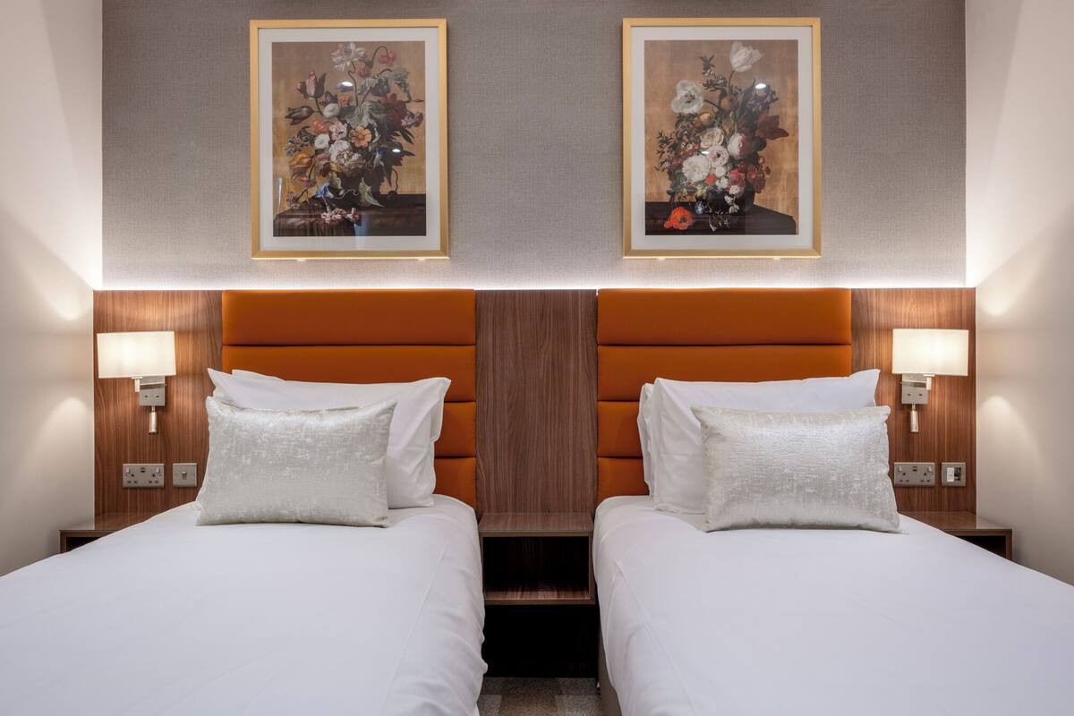 A hotel room with two beds, a wooden headboard, bedside lamps, and two framed floral paintings on the wall.