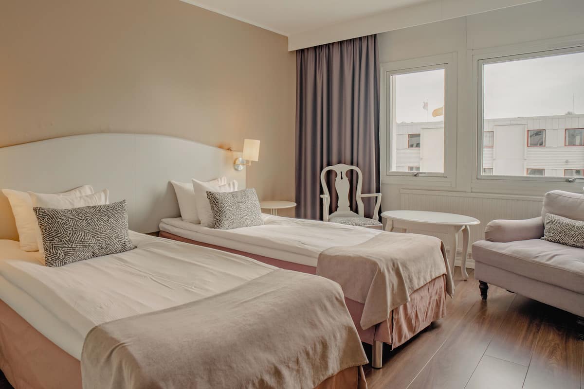Une chambre d’hôtel avec deux lits, une chaise, une petite table et une fenêtre donnant sur des immeubles.