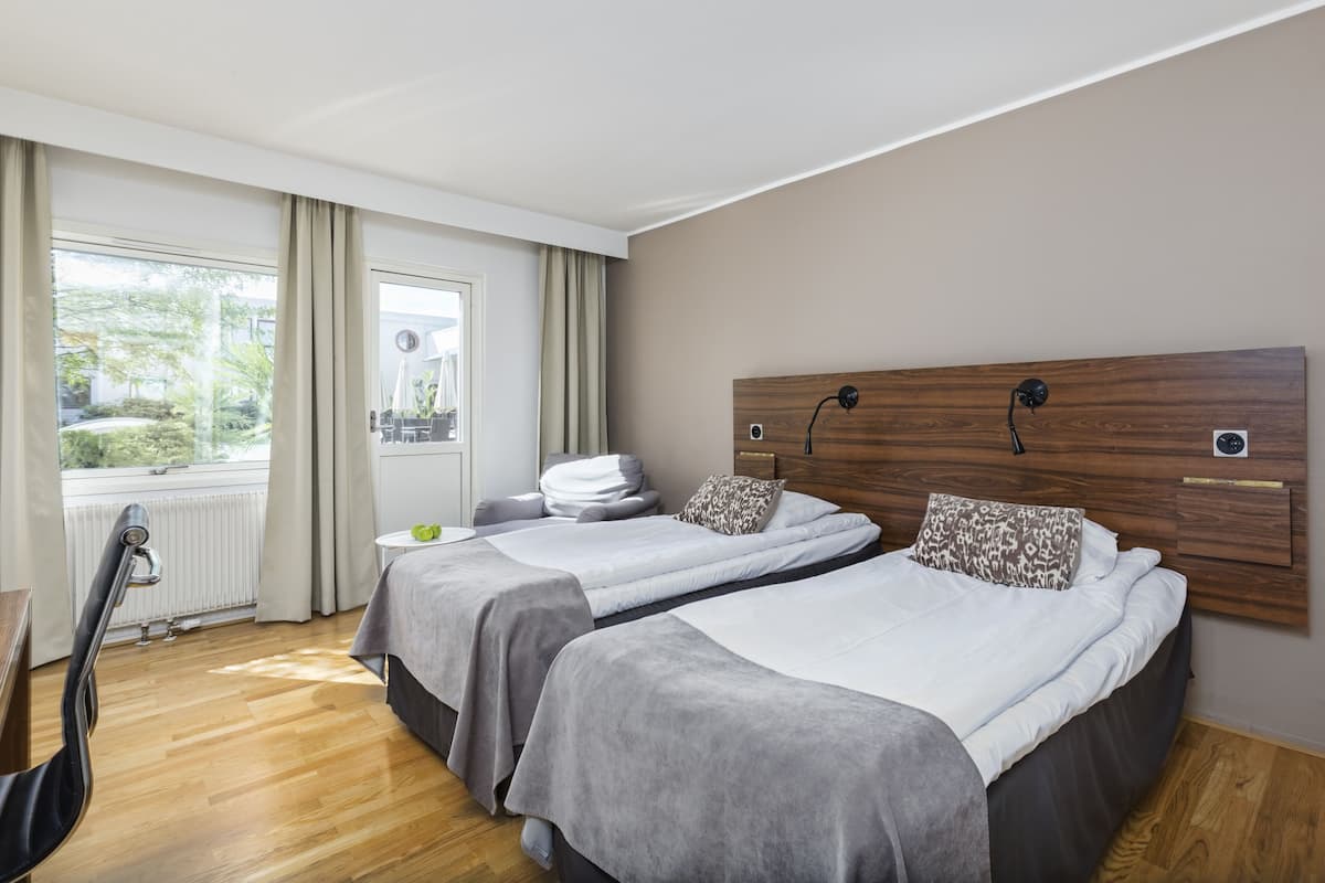 Une chambre d’hôtel avec deux lits, une vue sur l’extérieur, un radiateur et une petite table avec une plante.