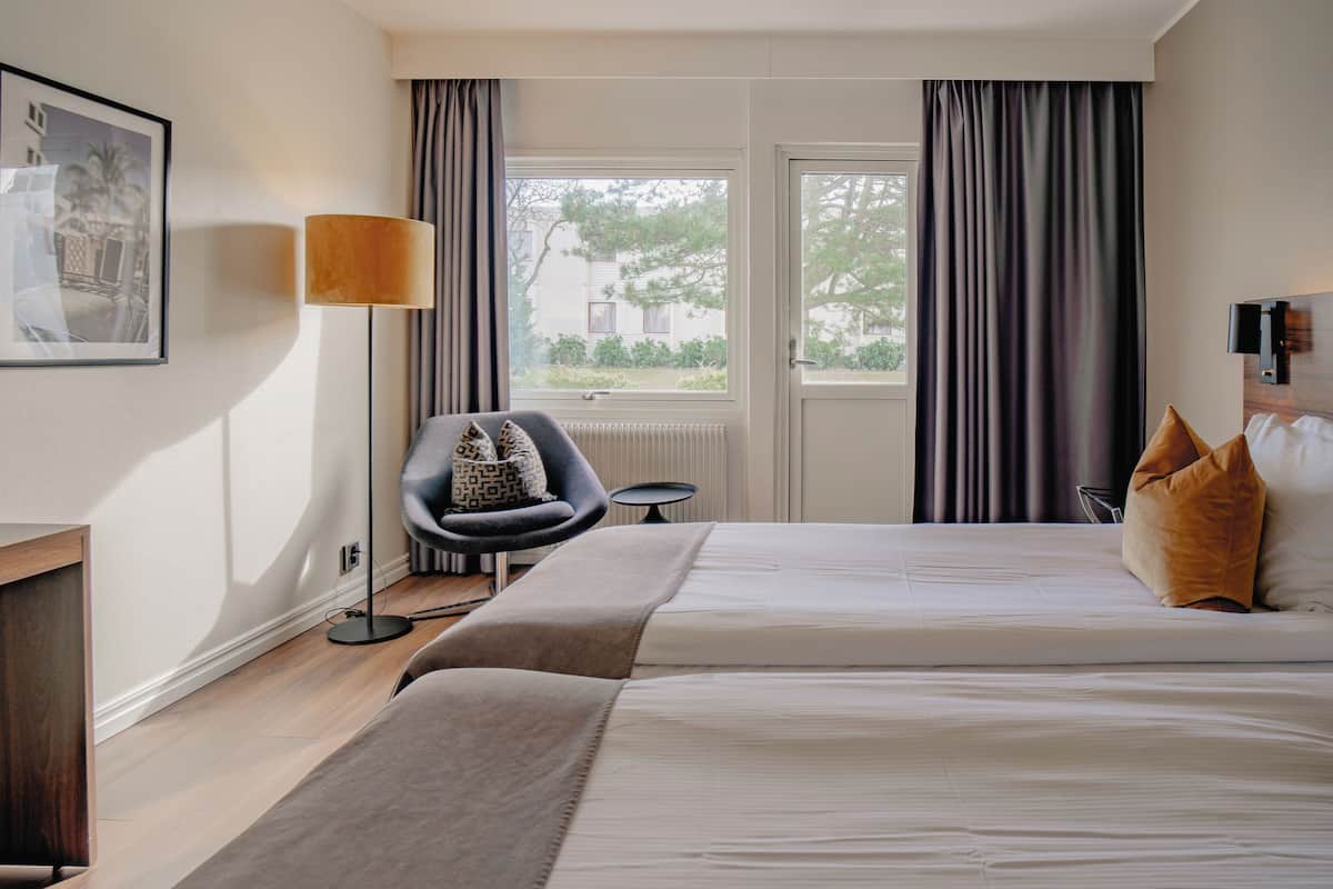 Une chambre d’hôtel avec un lit, un bureau, une chaise, une lampe et une fenêtre avec des rideaux.