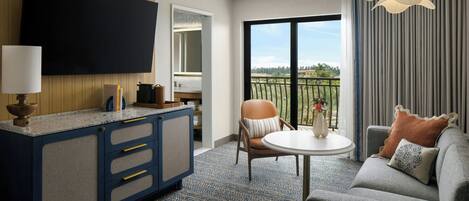 A hotel room with a flat-screen TV, a kitchenette, a sofa, a dining table, and a balcony with a view.