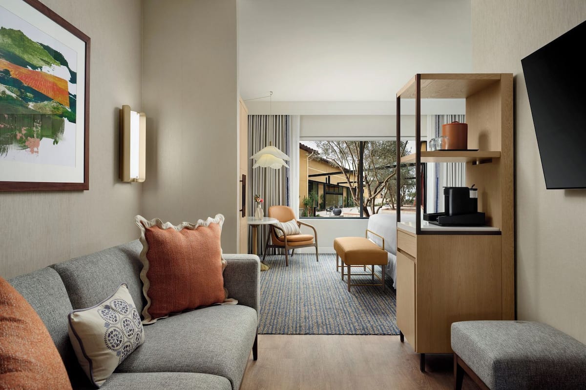 Ein modernes Hotelzimmer mit grauem Sofa, einem kleinen Tisch, einem Sessel, einer Holzkommode und einem großen Fenster mit Blick ins Freie.