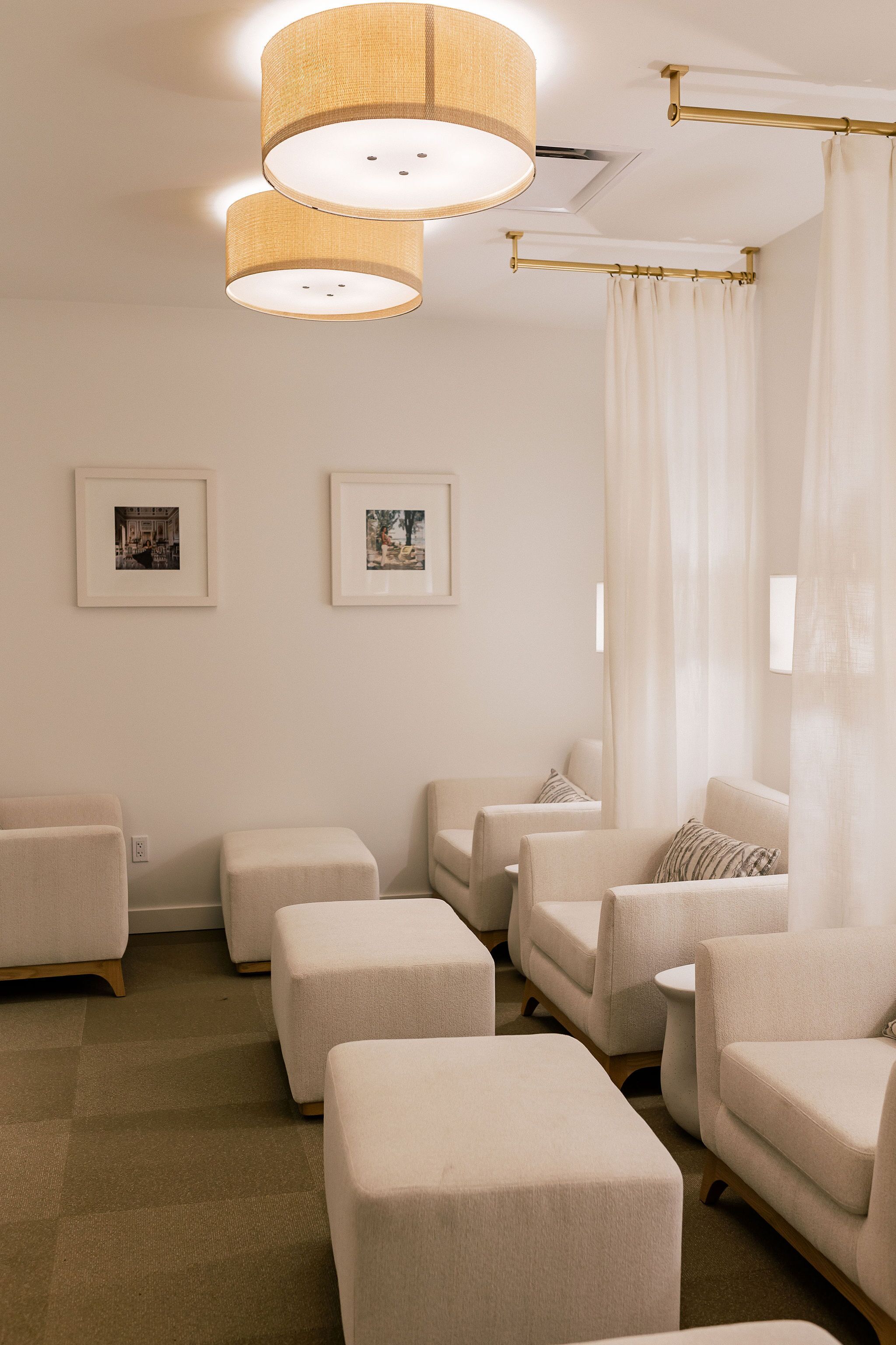 A modern hotel room with a neutral color palette, featuring a seating area with sofas and ottomans, a ceiling light fixture, and framed pictures on the wall.