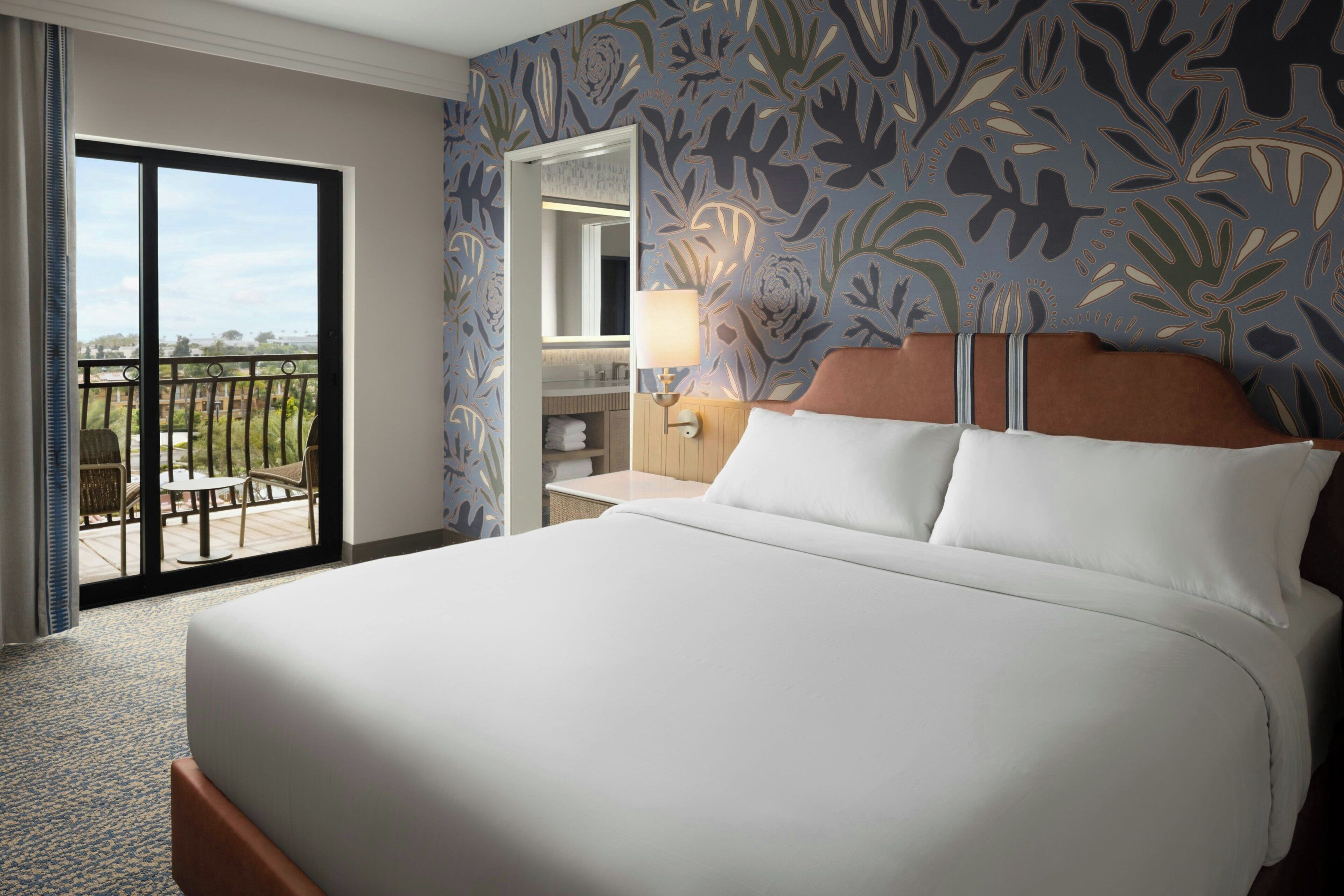 A hotel room with a large bed, a balcony with a view, a bathroom with a mirror, and a wall with a leaf pattern.