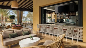 Un área de bar al aire libre con sofá y sillones, una cocina al fondo y vista a palmeras.