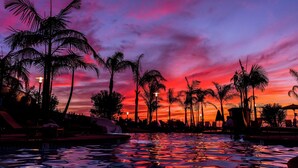 Un área de piscina con palmeras, sillas de descanso y un cielo al atardecer.