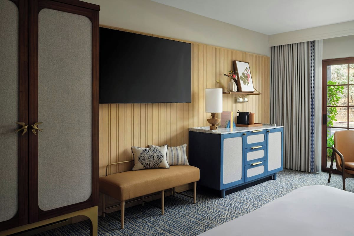 Ein Hotelzimmer mit einem Bett, einer Bank, einem Schreibtisch, einem Fernseher und einem Fenster mit Vorhängen.